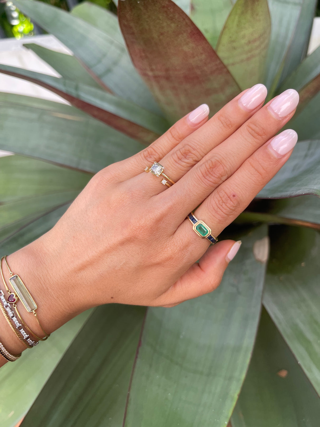 A gold ring with a central emerald flanked by two step-cut sapphires on either side, worn with diamond engagement ring with a green flower in the background.