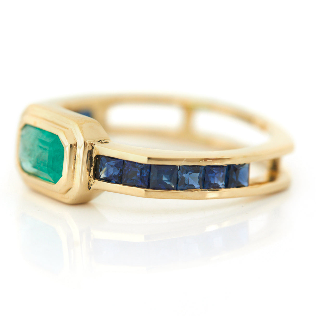 A gold ring with a central emerald flanked by many step-cut sapphires on either side.