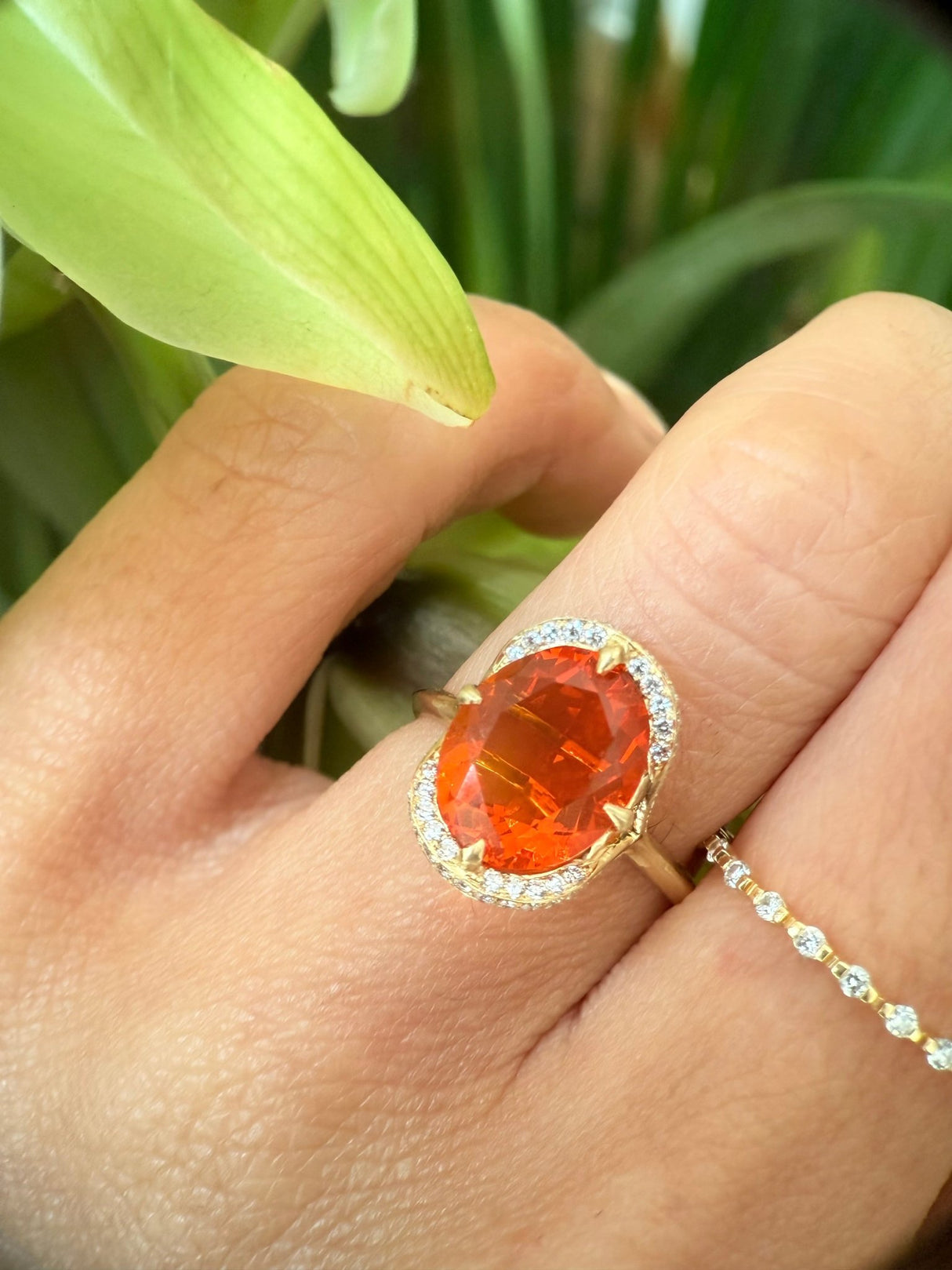 Close up of a Fire opal engagement ring in 18k gold worn on model against a leafy background