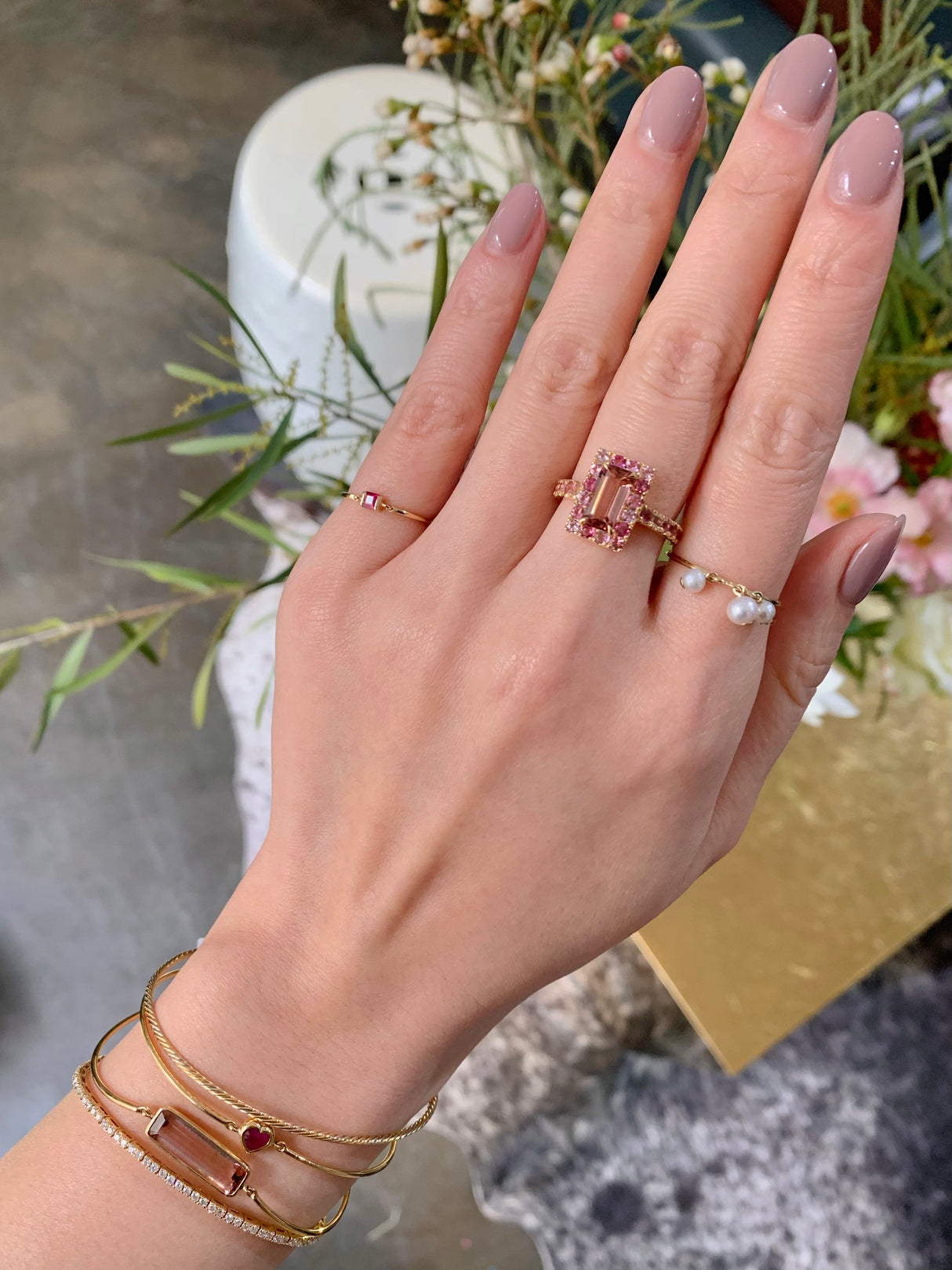An 18k yellow gold ring with a central baguette cut pink tourmaline surrounded by a frame of smaller round multi colored pink tourmalines worn on model. Ring stacked with other rings for a modern look. Perfect engagement ring or wedding ring.