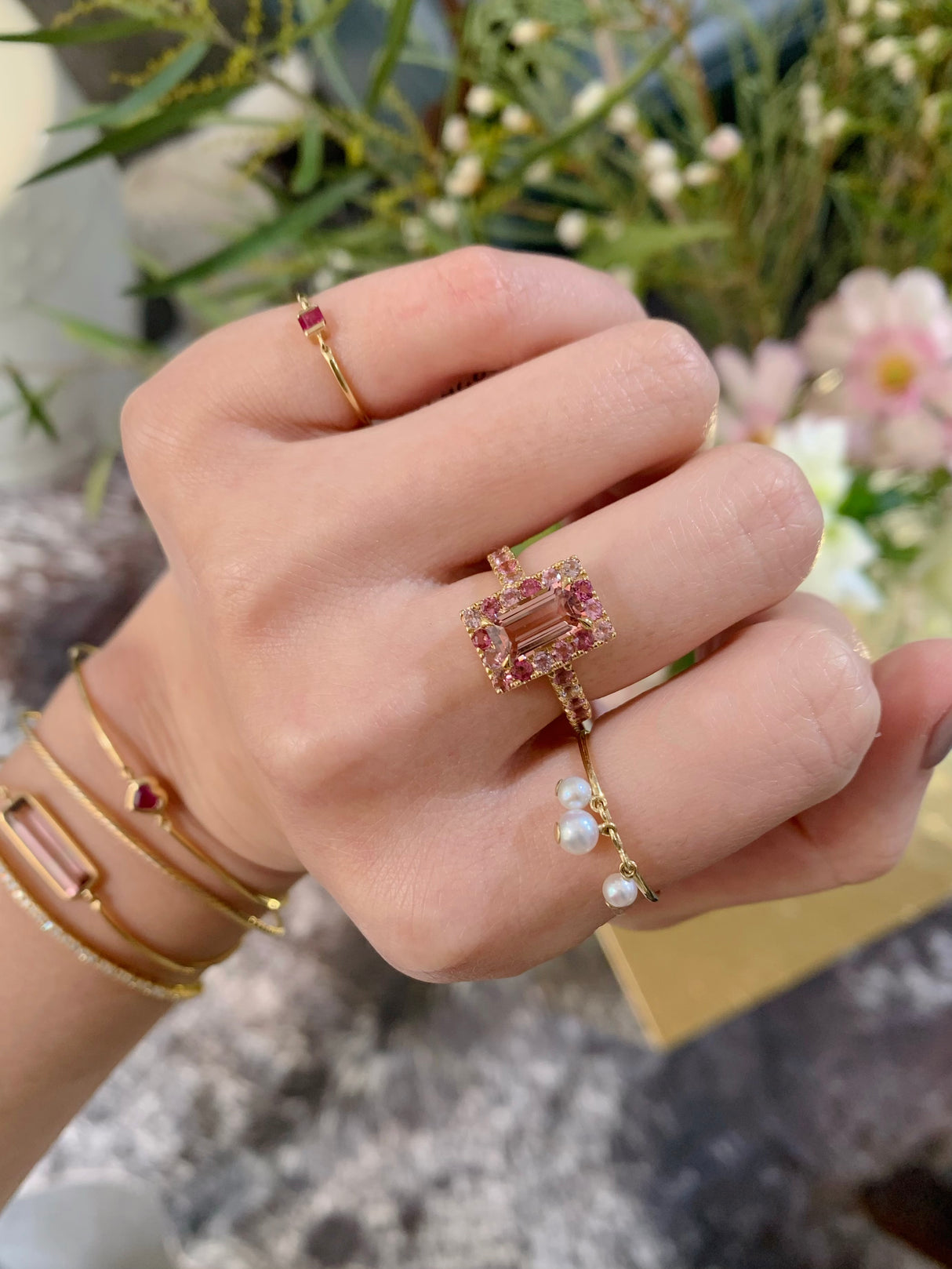 An 18k yellow gold ring with a central baguette cut pink tourmaline surrounded by a frame of smaller round multi colored pink tourmalines worn on model. Ring stacked with other rings for a modern look. Perfect engagement ring or wedding ring.