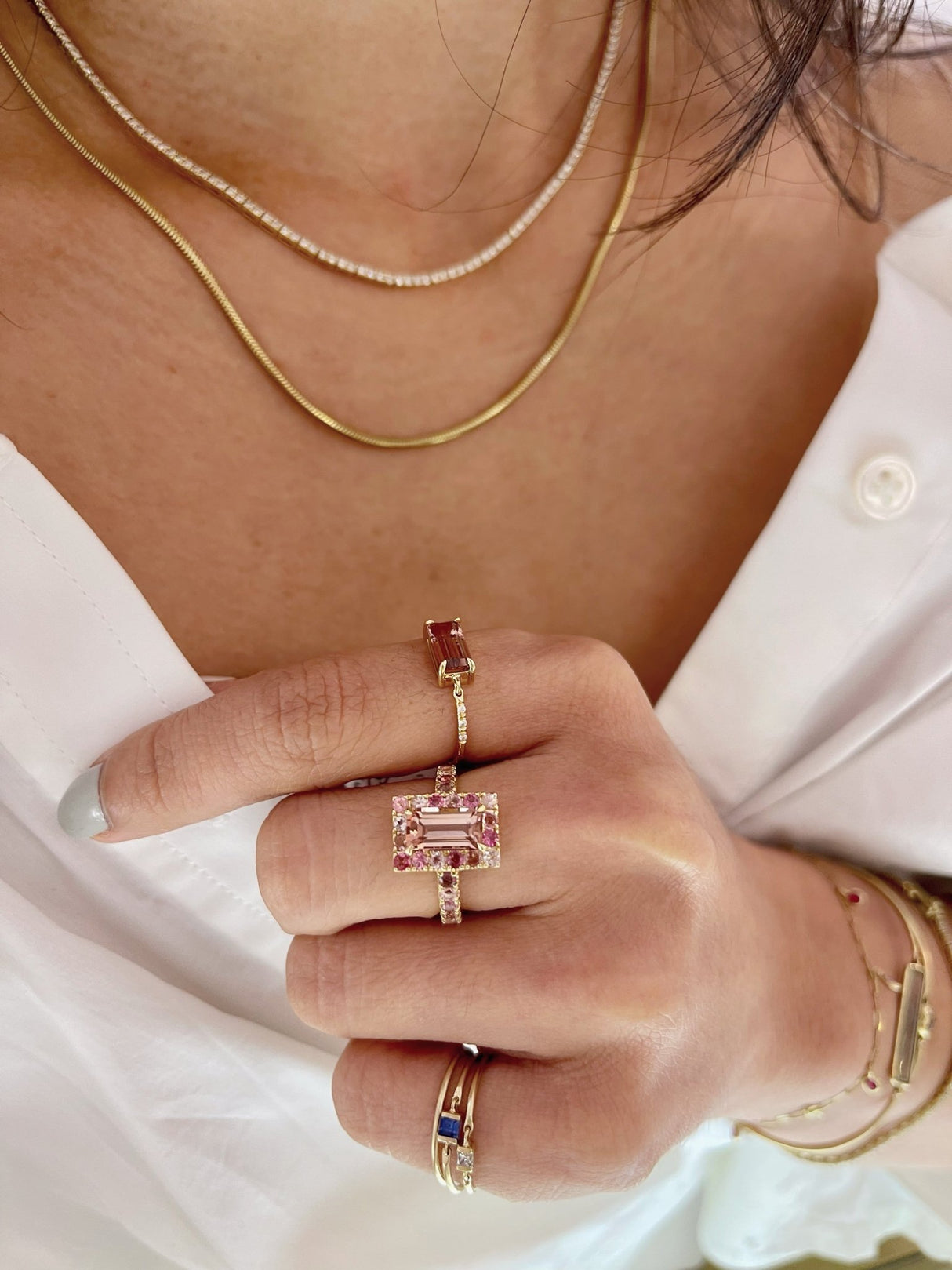An 18k yellow gold ring with a central baguette cut pink tourmaline surrounded by a frame of smaller round multi colored pink tourmalines worn on model. Ring stacked with other rings for a modern look. Perfect engagement ring or wedding ring.