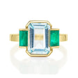 An 18K gold ring with a central aquamarine gem flanked by two vivid green emerald gems on the sides. 