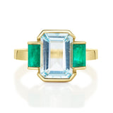 An 18K gold ring with a central aquamarine gem flanked by two vivid green emerald gems on the sides. 