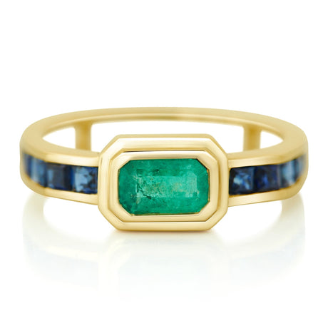 A gold ring with a central emerald flanked by many step-cut sapphires on either side, set against a white background.