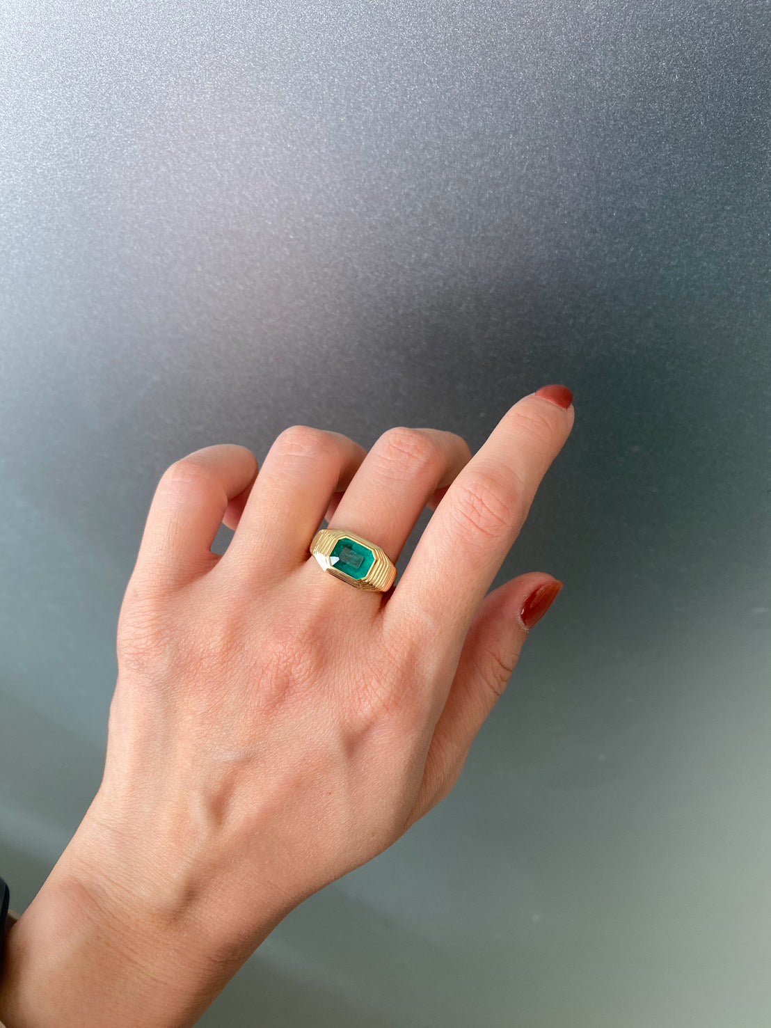 Hand wearing a gold ring with a large emerald against a plain background