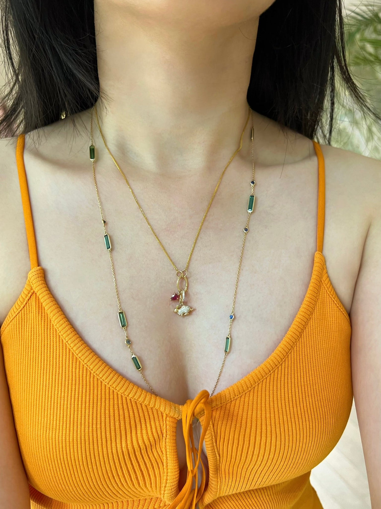 Person wearing a delicate necklace with green gemstones and a small pendant, against a blurred natural background.
